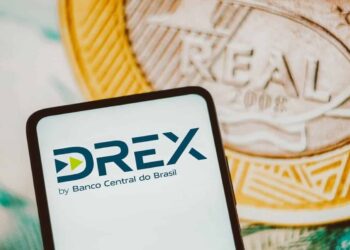 Tela de celular exibindo o logo do DREX, moeda digital do Banco Central do Brasil, com moedas ao fundo.