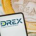 Tela de celular exibindo o logo do DREX, moeda digital do Banco Central do Brasil, com moedas ao fundo.