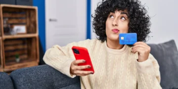 Mulher com cabelo cacheado segurando um cartão de crédito e um celular, com expressão pensativa.