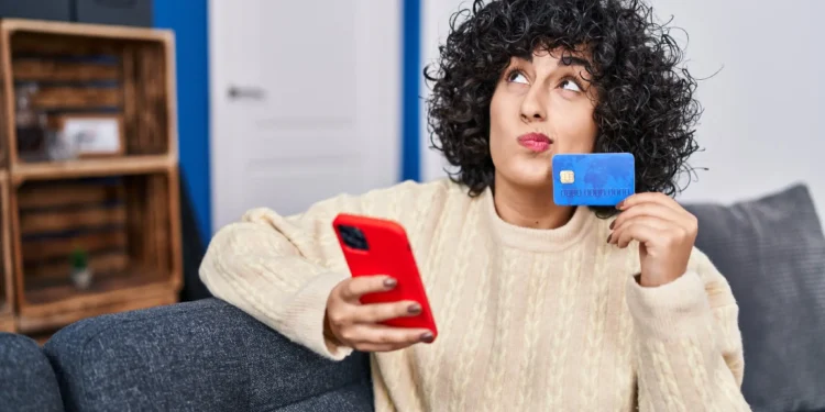 Mulher com cabelo cacheado segurando um cartão de crédito e um celular, com expressão pensativa.